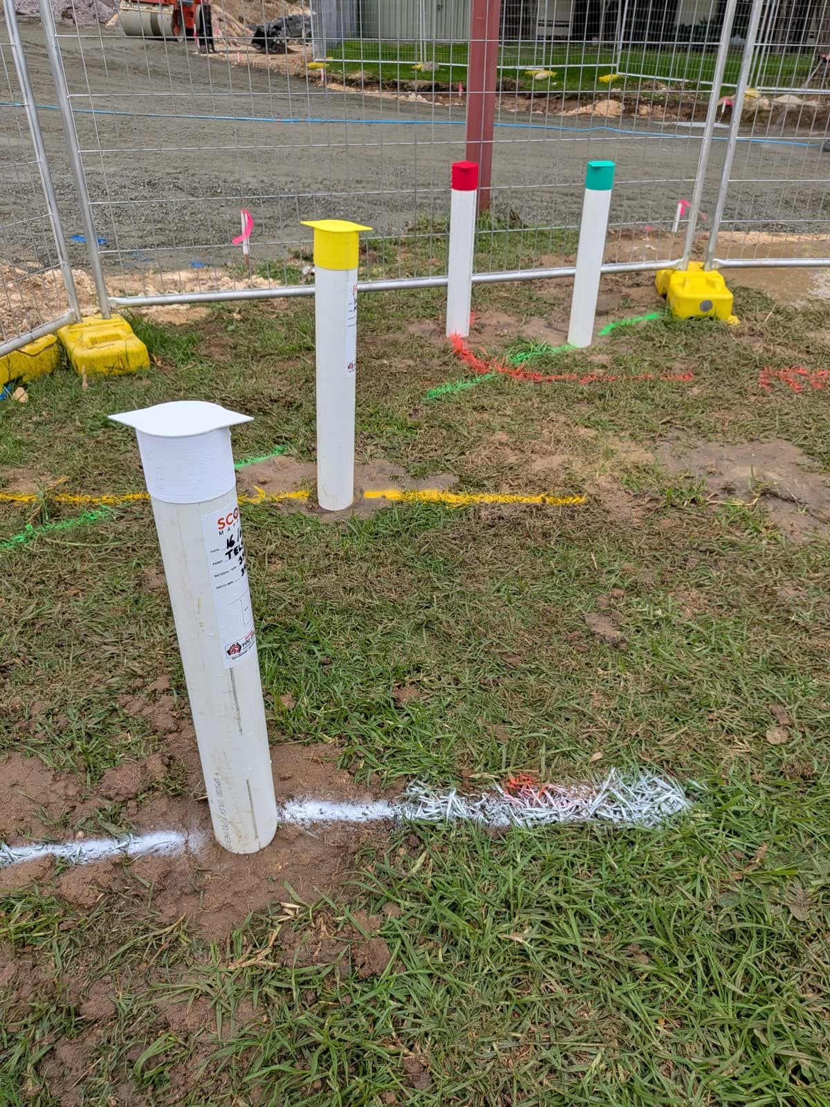 Underground services marked out on grass beside a construction site, with colour-capped marker posts and red, green, yellow and white spray lines